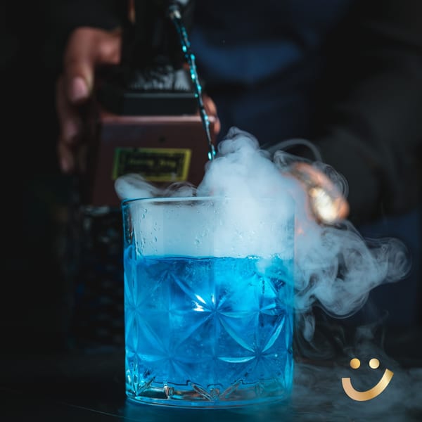 A blue cocktail in a textured glass with smoke billowing from the top, being poured by a bartender.