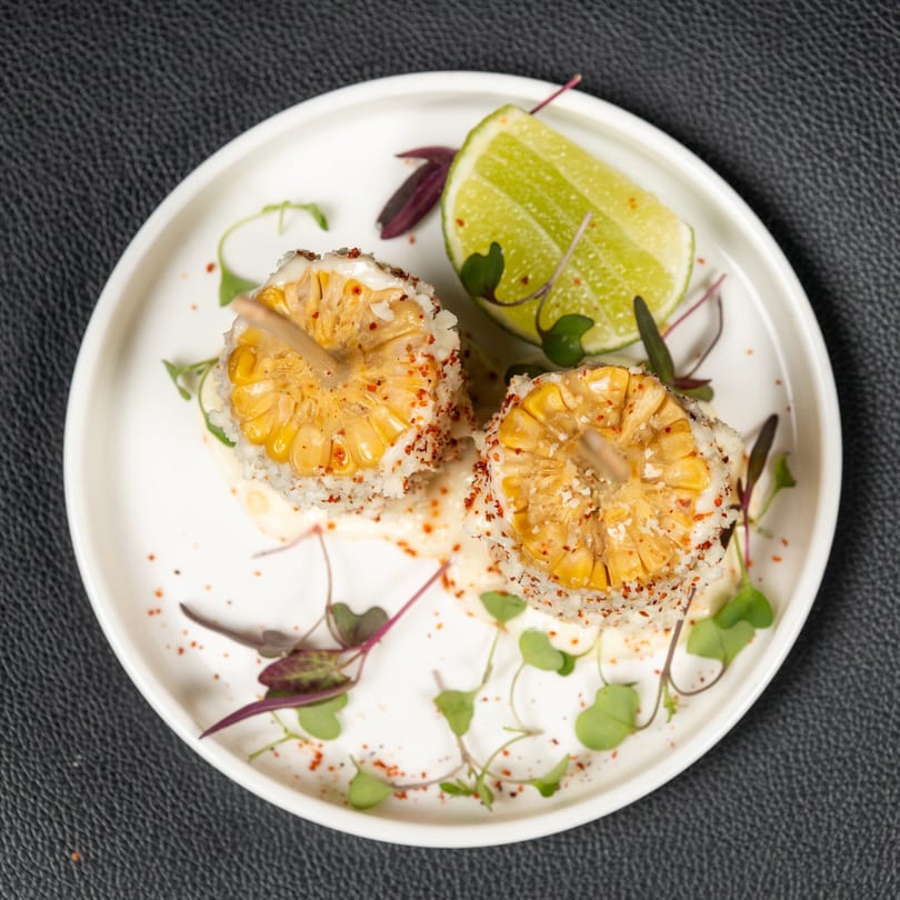 Two grilled corn on the cob pieces with white sauce, chili flakes, and a lime wedge.