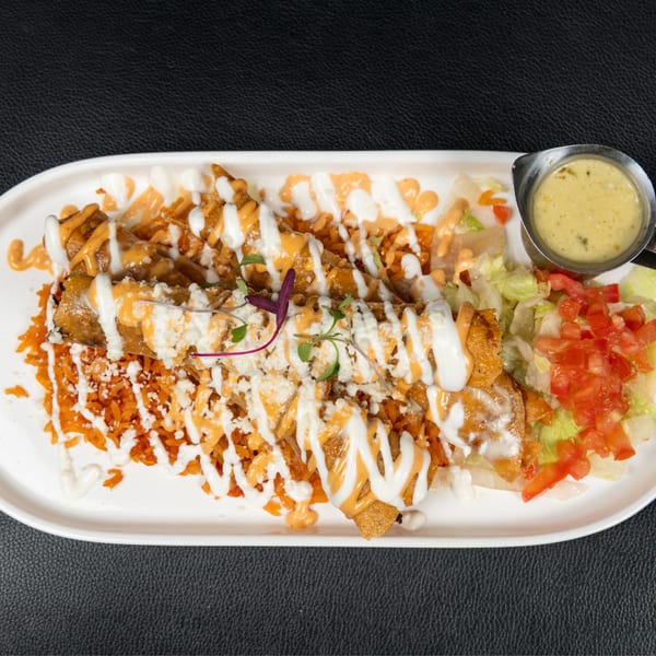 Crispy flautas drizzled with white and orange sauces, served with rice, salad, and a side of green sauce.