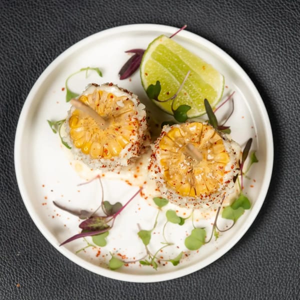 Two grilled corn on the cob halves with seasoning, sauce, and a lime wedge on a white plate.