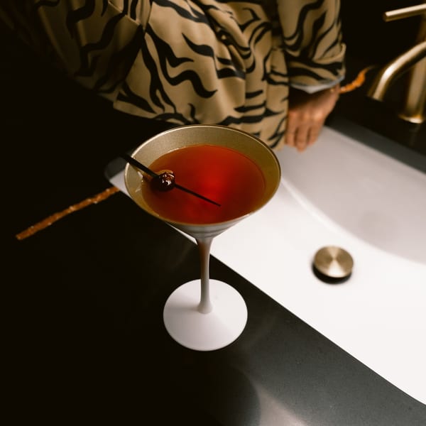 A red cocktail in a martini glass with a cherry, next to a sink with a gold faucet.