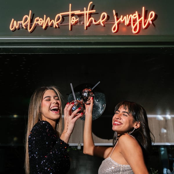 Two women holding disco ball drinks, laughing under a neon "welcome to the jungle" sign.