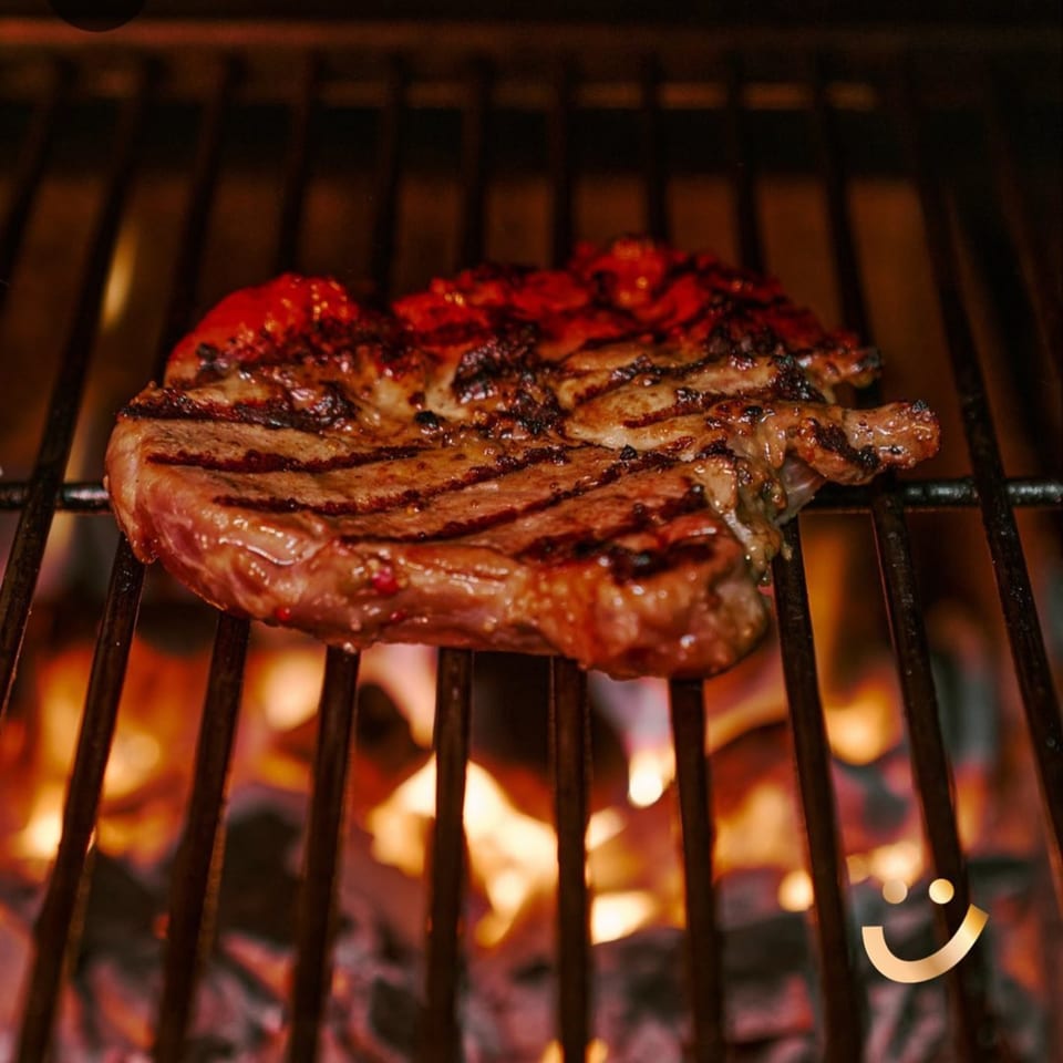 A sizzling steak with red sauce grilling over hot coals, ready to be served at La Buena Vida.