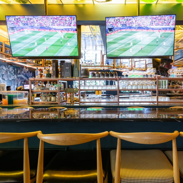 A bar with two large TVs showing a soccer game, bottles, glasses, and bar stools.