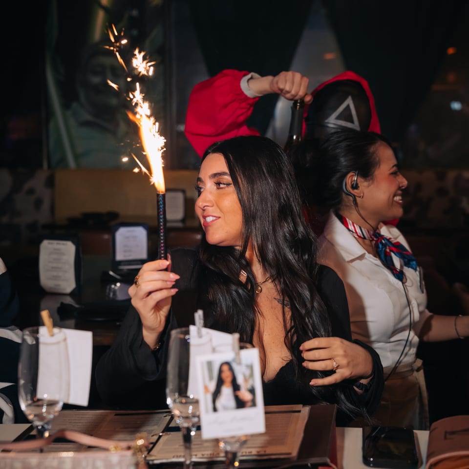 A woman holding a sparkling firework at a table, with a server and another person in the background.