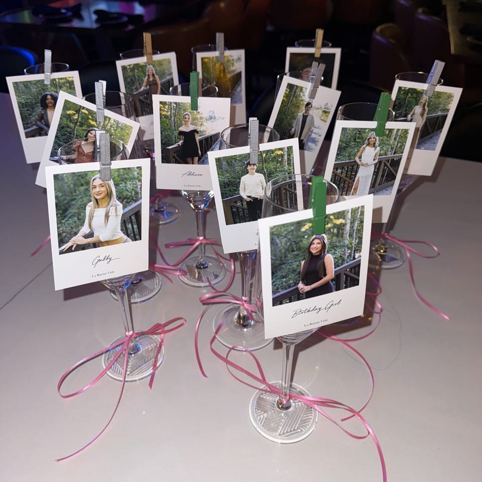 Wine glasses with personalized photos clipped to their rims, adorned with pink ribbons, on a white table.