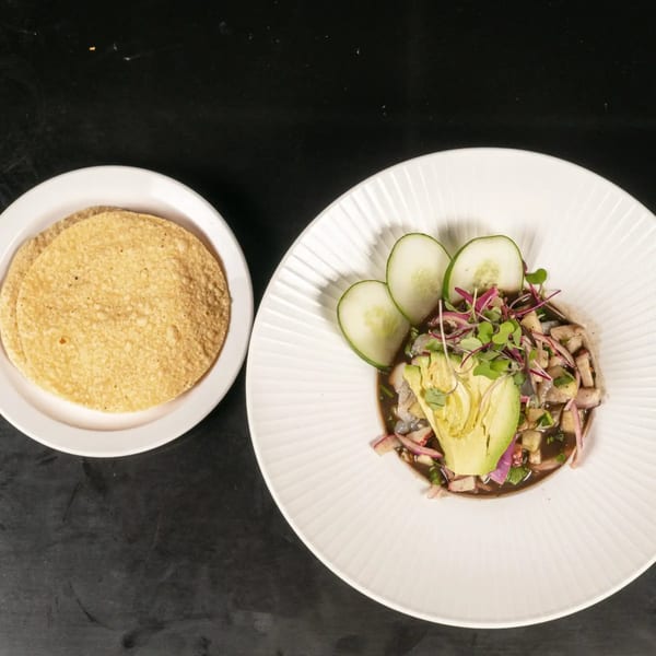 A white plate of ceviche with avocado, cucumber, and red onion, next to a plate of tortillas.