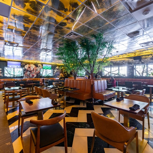 A restaurant interior with brown booths, wooden tables, black and white checkered floor, and artificial trees.