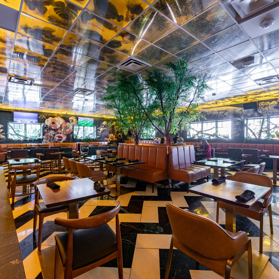 A restaurant interior with brown booths, wooden tables, black and white checkered floor, and artificial trees.