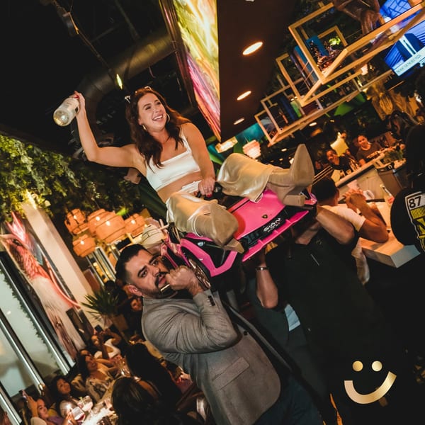 A woman on a pink toy car, held by men, cheers with a bottle in a lively La Buena Vida restaurant.