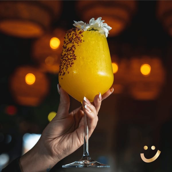 Hand holding a vibrant yellow La Buena Vida cocktail garnished with chili flakes and white flowers.