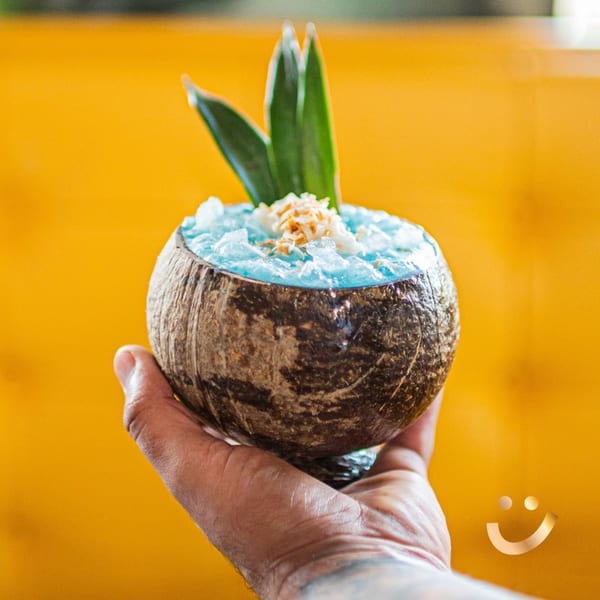 Hand holding a coconut shell filled with a blue drink, ice, and garnished with toasted coconut and leaves.