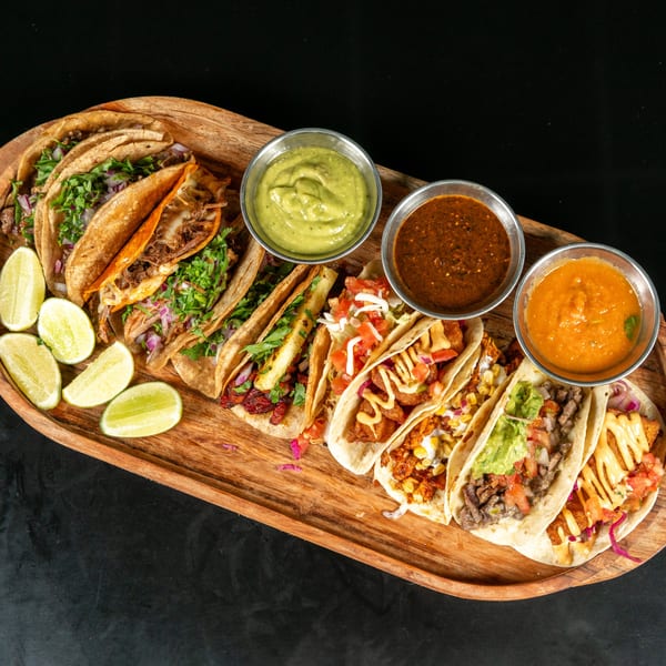 A wooden platter filled with various tacos, lime wedges, and three bowls of dipping sauces.
