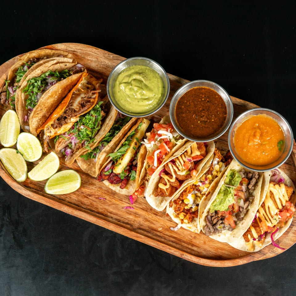 A wooden platter filled with various tacos, lime wedges, and three bowls of dipping sauces.