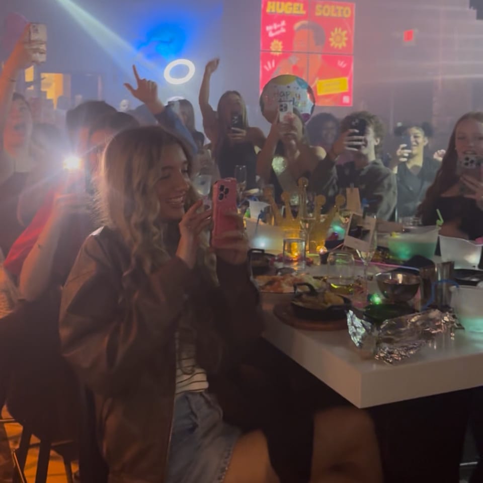People celebrating at La Buena Vida, with bright lights, phones recording, and drinks on a table.