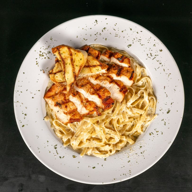 Grilled chicken and garlic bread served over creamy fettuccine alfredo on a white plate.
