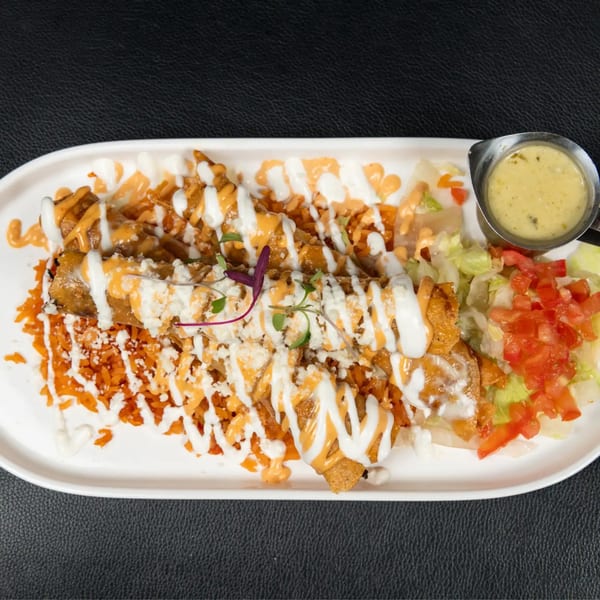 Crispy taquitos drizzled with white and orange sauces, served with red rice, salad, and a side of green sauce.