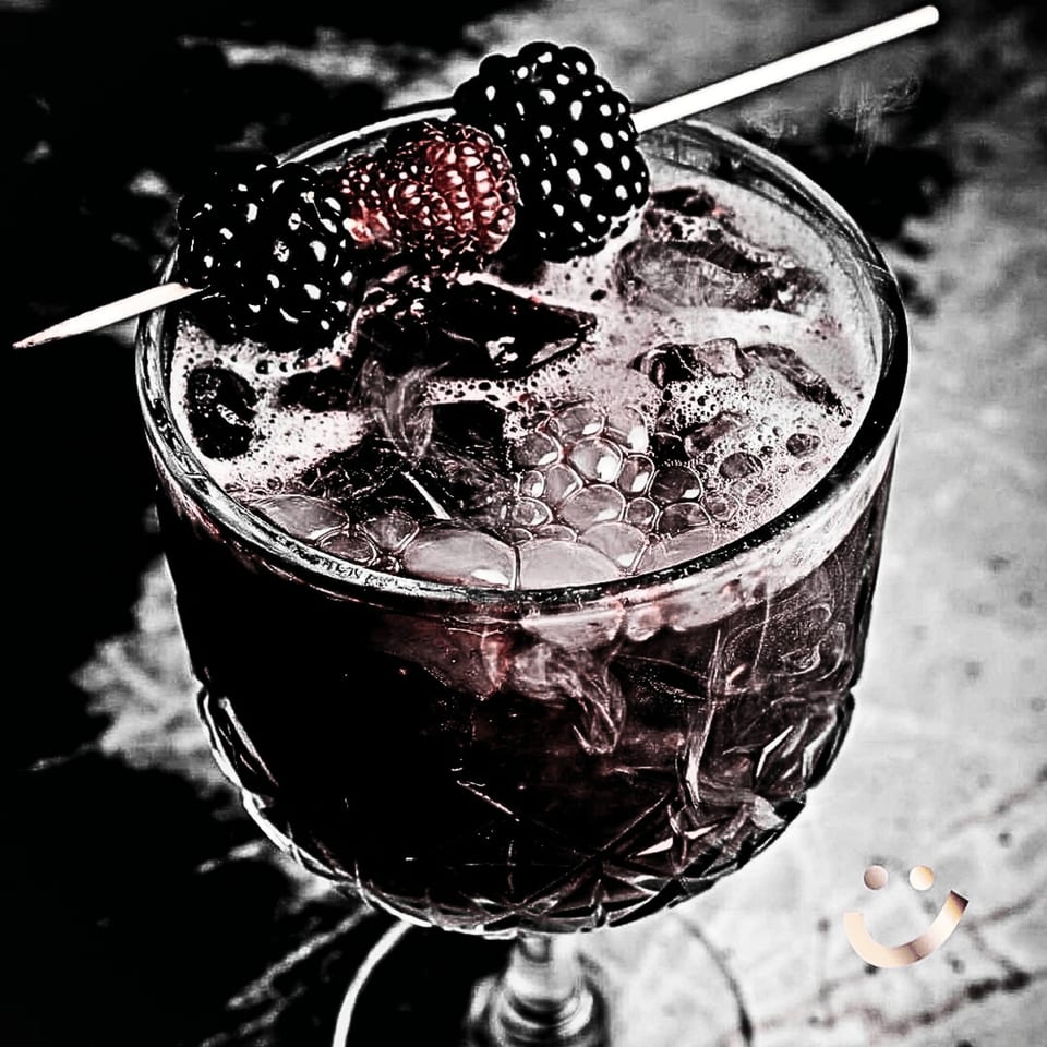 Dark berry cocktail with ice and foam, garnished with blackberries and raspberries on a skewer.