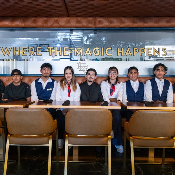 Six restaurant staff members in uniform sit at tables with a "WHERE THE MAGIC HAPPENS" sign behind them.