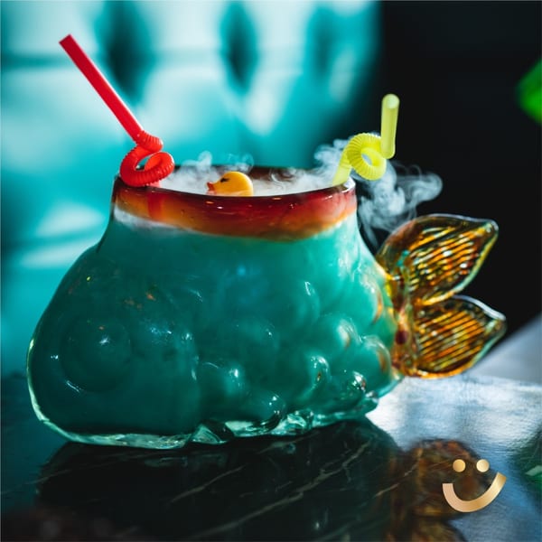A smoking blue cocktail in a fish-shaped glass with a rubber duck and two straws.