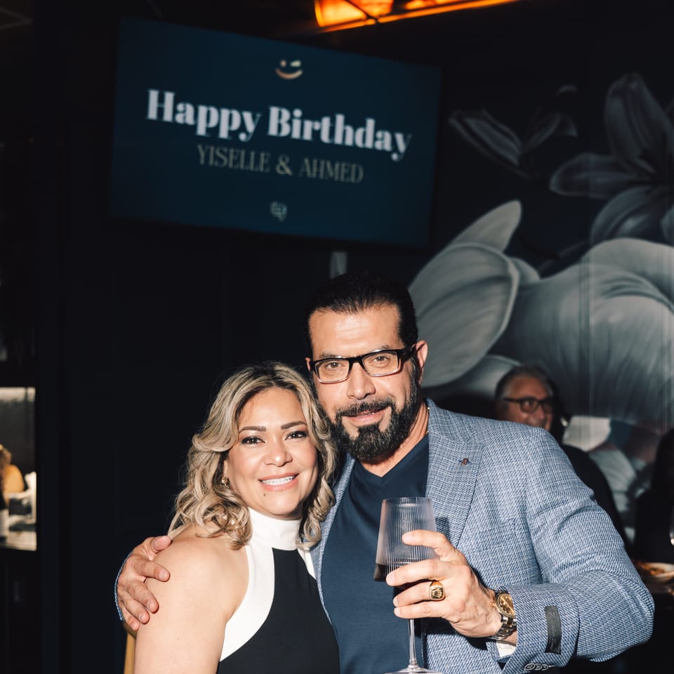 A smiling couple poses for a photo, with a "Happy Birthday Yiselle & Ahmed" sign in the background.