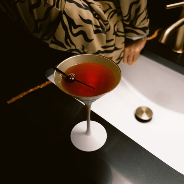 A red cocktail in a martini glass with a cherry, next to a sink with a gold faucet.