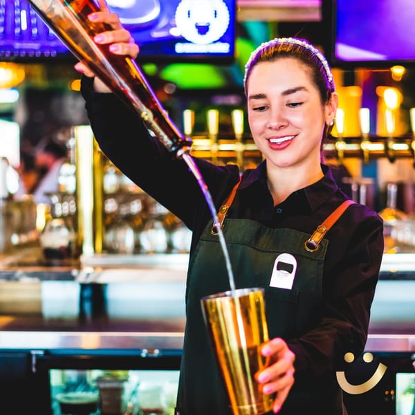 A smiling La Buena Vida bartender pours a drink from a bottle into a golden shaker.