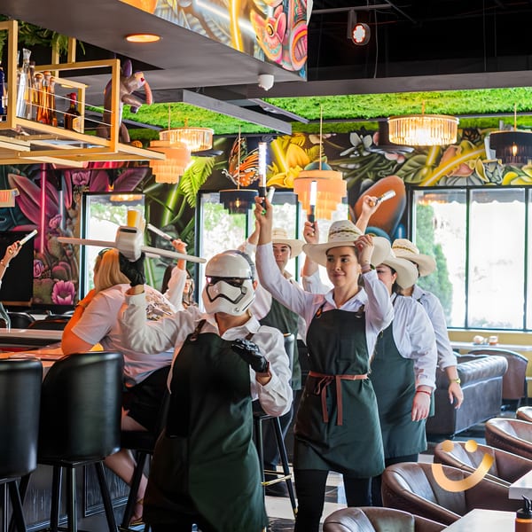 La Buena Vida staff in aprons and hats, one in a Stormtrooper helmet, raising light-up sticks.