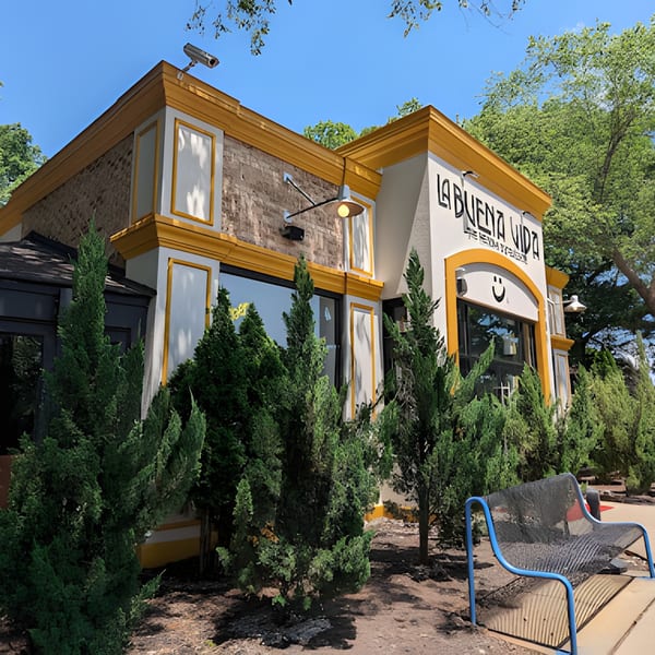 Exterior of La Buena Vida Mexican restaurant with yellow trim, surrounded by green shrubs and trees.