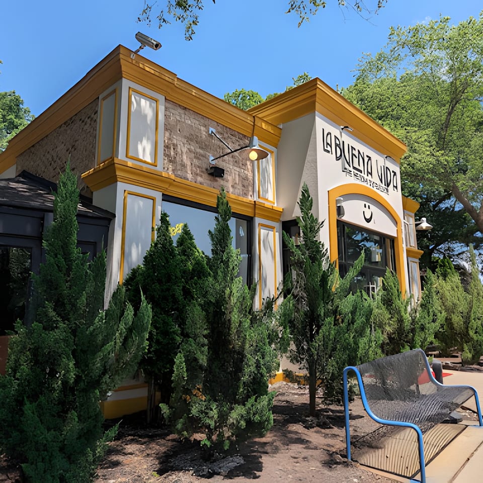 Exterior of La Buena Vida Mexican restaurant with yellow trim, surrounded by green shrubs and trees.