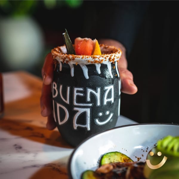 A hand holding a black La Buena Vida branded mug with a garnished, rimmed drink.