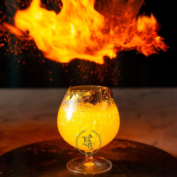 A La Buena Vida cocktail with gold leaf, glowing orange, with flames and sparks above it.