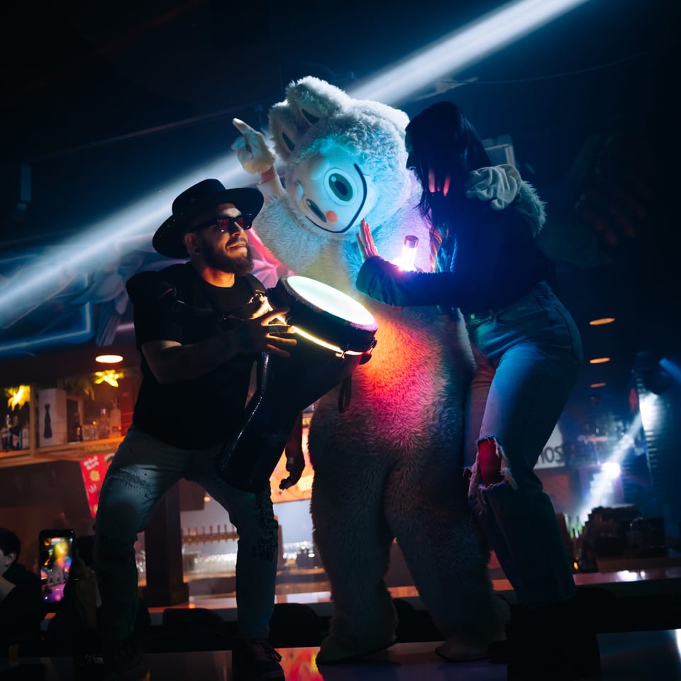 A man playing a drum, a woman, and a mascot dancing under blue and pink lights at La Buena Vida.